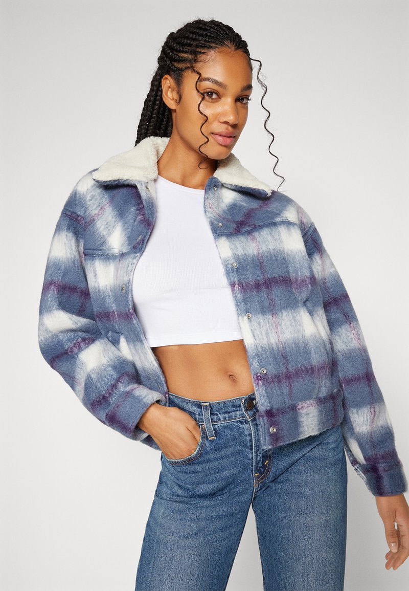 Veste en flanelle à carreaux bleu et blanc avec des accents violets, dotée d'un col en fausse fourrure et d'une fermeture à boutons-pression. Portée sur un crop top blanc.