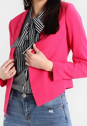 Frau trägt einen hellpinkfarbenen Blazer über einer schwarz-weiß gestreiften Bluse mit Schleife, kombiniert mit blauen Jeans.