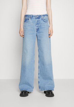 Jeans de pierna ancha en azul claro con un acabado desgastado, que presentan un diseño clásico de cinco bolsillos y un dobladillo sin terminar. Combinados con zapatos negros de suela gruesa.