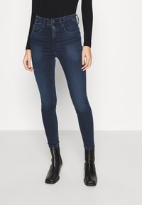 Mujer con jeans skinny de talle alto azul oscuro, blusa de manga larga negra con textura en canalé y botas negras hasta el tobillo, de pie contra un fondo blanco.