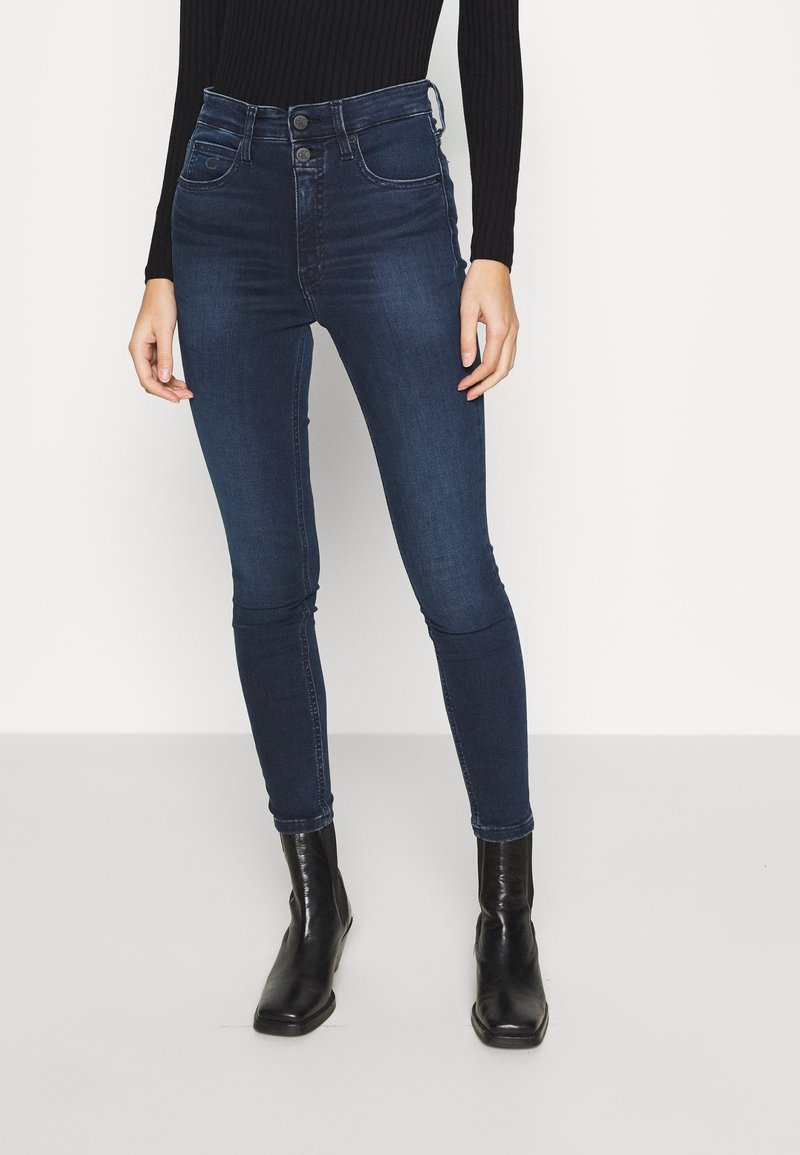 Mujer con jeans skinny de talle alto azul oscuro, blusa de manga larga negra con textura en canalé y botas negras hasta el tobillo, de pie contra un fondo blanco.