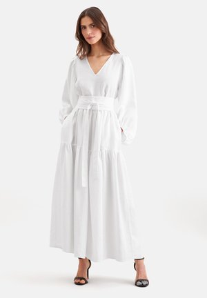 Femme portant une longue robe blanche à col en V, manches longues bouffantes, large ceinture à la taille et talons noirs ouverts à l'avant, debout devant un fond blanc.
