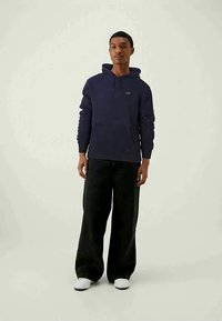 Jovem em pé, vestindo um hoodie azul-marinho, calças largas pretas e ténis brancos, contra um fundo claro e simples.
