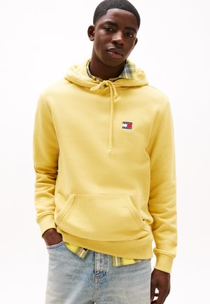 Sudadera con capucha amarilla hecha de una tela suave, que presenta un bolsillo frontal y un parche de logo. Superpuesta a una camisa de cuadros, combinada con jeans claros.