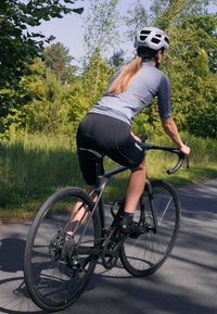 Schwarzes Rennrad mit sleekem Design, leichtem Rahmen und schmalen Reifen. Der Fahrer trägt ein graues T-Shirt und schwarze Shorts und befindet sich auf einem gepflasterten Weg.