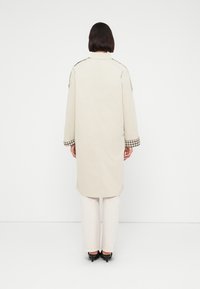 Μπεζ μακρύ παλτό με επωμίδες στους ώμους και μανσέτες με καρό σχέδιο. Φορεμένο πάνω από λευκό παντελόνι, με ίσια σιλουέτα και μίνιμαλ σχεδιασμό.