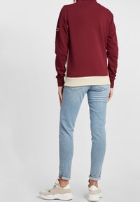 Personne portant un pull bordeaux et beige, un jean skinny bleu clair retroussé et des baskets beige et blanches, debout devant un fond uni.