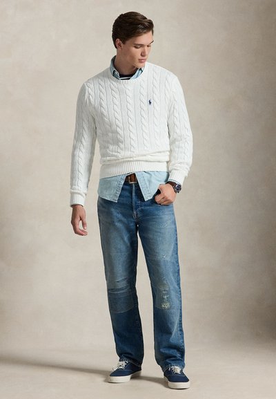 Jeune homme portant un pull en maille torsadée blanc par-dessus une chemise en denim bleu, avec un jean bleu et des baskets bleu marine, regardant vers le bas à sa gauche.