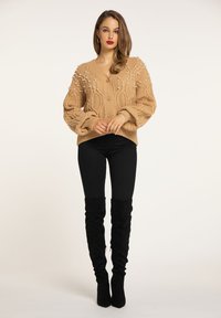 Pulover bej tricotat cu accente de perle 3D, decolteu în V și model cu cabluri; asociat cu blugi negri skinny și cizme negre până la coapsă.