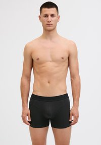 Young man standing facing forward, wearing black boxer briefs with "JACK & JONES" on the waistband, plain light background.