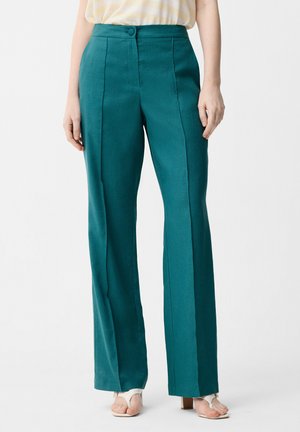 Femme portant un pantalon taille haute ajusté de couleur sarcelle avec un pli à l'avant et des sandales blanches à talons ouverts, debout devant un fond uni.