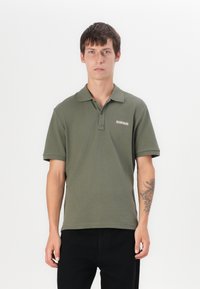 Oliven grønn polo skjorte med klassisk krage og kortermede. Har et logo på brystet og en glatt bomullsoverflate. Kombinert med svarte bukser.