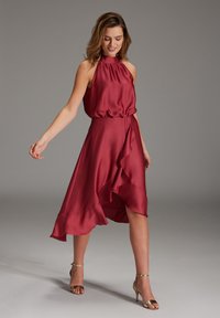 Robe en satin bourgogne dotée d'un col halter, de fronces à la taille et d'un ourlet asymétrique à volants. Des sandales à talons hauts dorées viennent parfaire le look.
