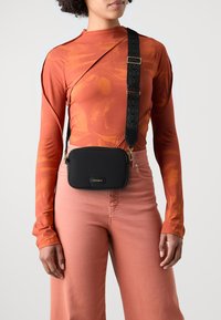 Borsa a tracolla nera in nylon con una tracolla regolabile a fantasia, dotata di una fibbia in metallo dorato e un logo sul davanti.