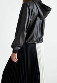 Giacca nera in raso con cappuccio, orlo elasticizzato e gonna plissettata con righe verticali in bianco e nero. Dettagli in oro sottile sui polsini.