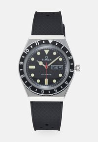Timex quartz watch with a black dial, silver stainless steel case, black textured rubber strap, and red second hand with date feature.