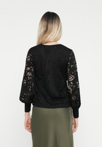 Μαύρη δαντελένια μπλούζα με μακριά, φουσκωτά μανίκια, πολύπλοκα λουλουδάτα μοτίβα και μονόχρωμο μαύρο ύφασμα στην πλάτη. Συνδυάζεται με πράσινη φούστα.