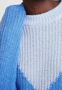 Pull en maille présentant une palette de couleurs bleu clair et blanc, avec une surface texturée et un motif en chevron au niveau de la poitrine.