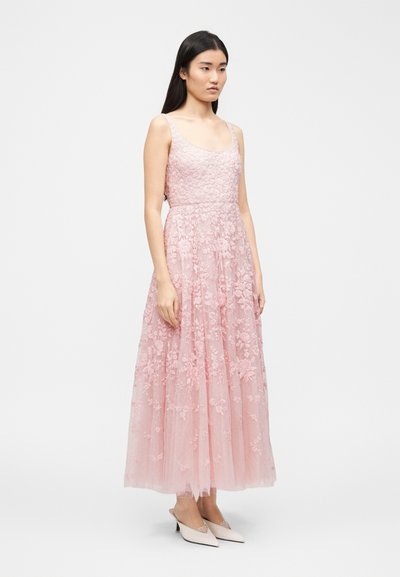 Νεαρή γυναίκα όρθια, φορώντας αμάνικο, μακρύ φόρεμα με ροζ λουλουδάτο δαντέλα και λευκές μυτερές γόβες kitten heels, σε λευκό φόντο.