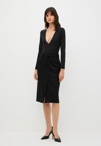 Musta pitkähiha bodysuit syvällä V-pääntiellä, yhdistettynä tiukkaan mustaan kynähameeseen, jossa on etu halkio ja vyöllinen vyötärö. Mustat korot.