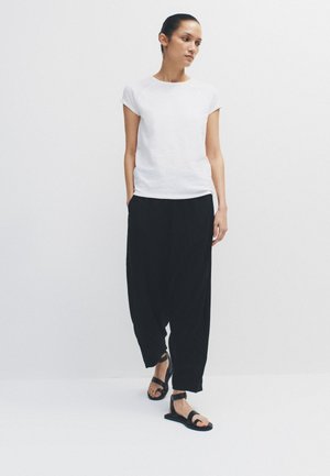 Vrouw in een witte top met korte mouwen, losse zwarte broek en zwarte sandalen met enkelbandje, lopend tegen een effen witte achtergrond.