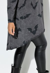 Ingrandimento di una persona che indossa un vestito grigio con stampe di pipistrello nero, leggings in ecopelle nera e stivali alla caviglia neri con borchie.