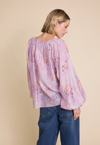 Lys lilla blomstret bluse med rynket halslinning og vide ærmer, parret med højtaljede blå denimjeans. Subtil struktureret stof.
