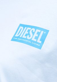 Λευκό ύφασμα με μπλε ορθογώνια ετικέτα που εμφανίζει το κείμενο "DIESEL FOR SUCCESSFUL LIVING" με έντονα και κεφαλαία γράμματα.