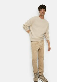 Man wearing beige ribbed sweater, light brown corduroy pants, and beige sneakers standing against a white background, posing casually.