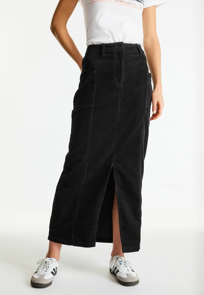 Femme portant un t-shirt blanc, une jupe midi en velours côtelé noire avec une fente à l'avant, et des baskets blanches avec des bandes noires, debout sur fond blanc.