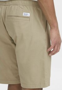 Beige shortsit, joissa on joustava vyötärö ja takatasku. Kankaan rakenne näyttää sileältä, ja lyhyt lahkeen pituus sekä vyötäröllä oleva etiketti lisätään.