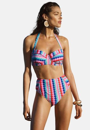 Vrouw die een kleurrijke bikini met hoge taille en een geschulpte print draagt, grote ronde oorbellen en gouden armbanden, staand tegen een witte achtergrond.