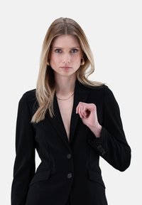 Schwarzer maßgeschneiderter Blazer aus strukturiertem Stoff, mit scharfen Revers, zwei Knöpfen und Seitentaschen. Modell trägt ihn und hält den Kragen leicht.