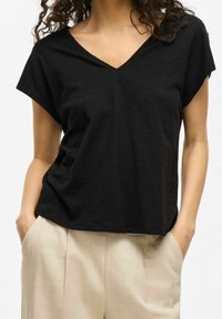 Haut noir à col en V avec manches courtes, en tissu texturé. Associé à un pantalon beige clair, mains dans les poches. Design minimaliste.