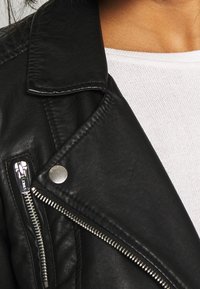 Närbild av en svart läderjacka med silverblixtlås och tryckknapp över en vit stickad topp på en persons övre bröst.