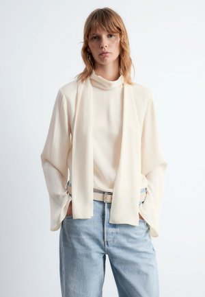 Cremfarbene Bluse mit drapiertem Ausschnitt und weiten Ärmeln, kombiniert mit hellblauen, hoch taillierten Jeans und einem beigen Gürtel.