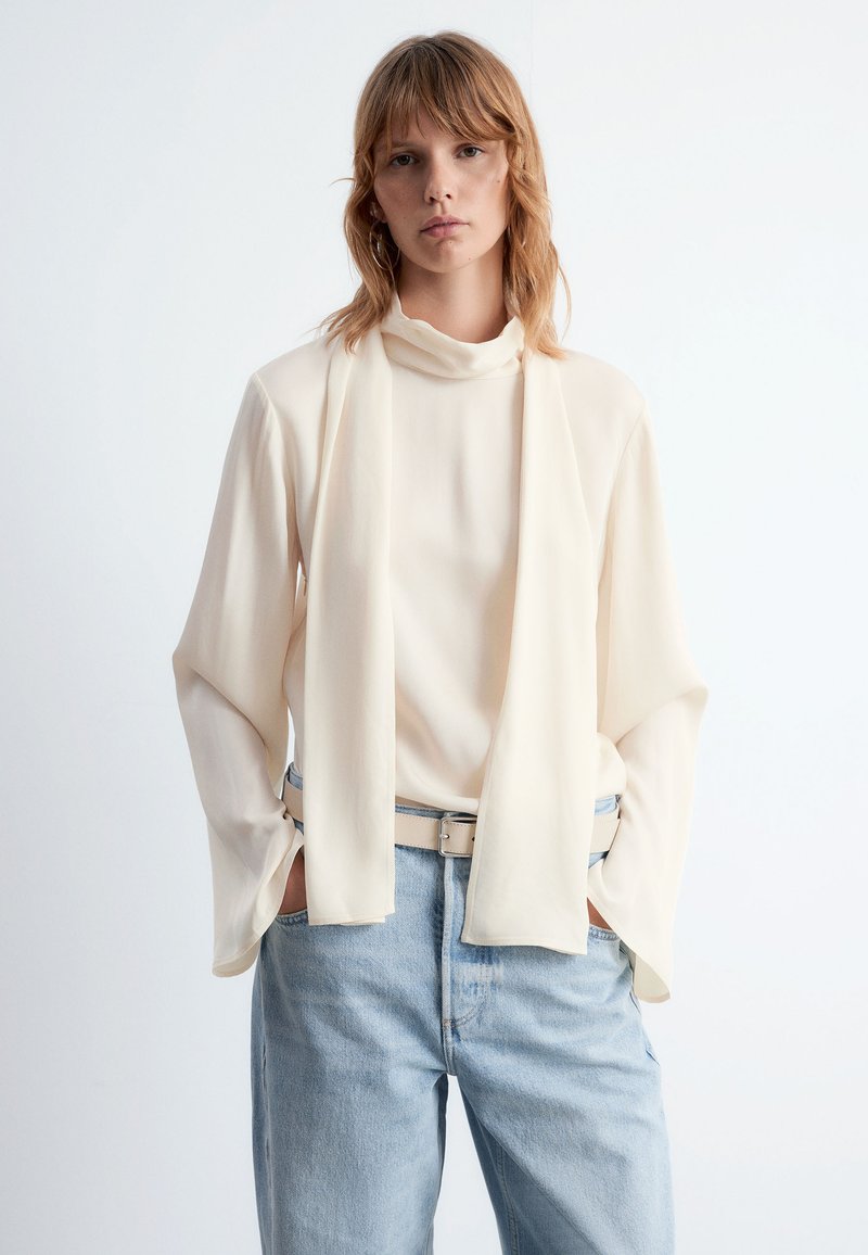 Cremfarbene Bluse mit drapiertem Ausschnitt und weiten Ärmeln, kombiniert mit hellblauen, hoch taillierten Jeans und einem beigen Gürtel.