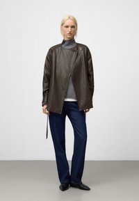Giacca di pelle marrone con colletto a revers, taglio asimmetrico e dettagli con bottoni. Abbinata a un dolcevita grigio e jeans blu scuro.