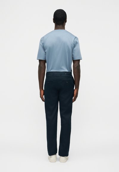 Άνδρας που στέκεται με την πλάτη στην κάμερα, φορώντας ανοιχτό μπλε κοντομάνικο πουκάμισο, σκούρο μπλε παντελόνι και λευκά αθλητικά σε λευκό φόντο.