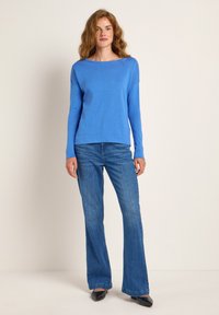 Blauer Langarmpullover mit Bootsausschnitt, kombiniert mit ausgestellten Jeans und schwarzen spitzen Schuhen. Glatte Textur, einfarbig, lockere Passform.