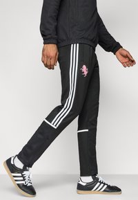 Μαύρο αθλητικό παντελόνι με λευκές ρίγες, διακοσμημένο με ροζ κεντητό λογότυπο μονόκερου. Φορεμένο με μαύρα αθλητικά παπούτσια, που αναδεικνύουν υφή υλικού.