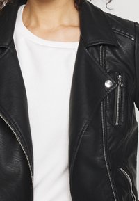 Svart lädermotorcykeljacka med silverfärgad dragkedja, tryckknappar och kviltade axeldetaljer, i kombination med en vit T-shirt med rund halsringning.