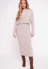 Kobieta w beżowym dzianinowym swetrze i dopasowanej długiej spódnicy oraz brązowych zamszowych kozakach za kolano, stojąca na białym tle.