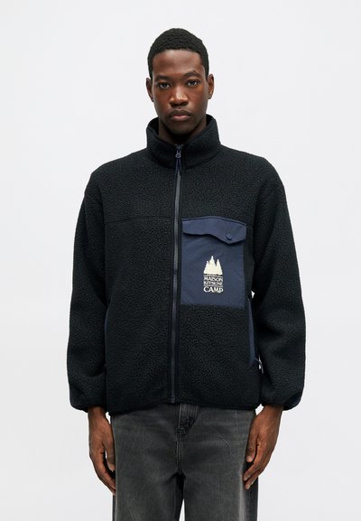 Μαύρο φleece μπουφάν με ναυτική τσέπη, διαθέτει κλείσιμο με φερμουάρ και λογότυπο σε χρυσό. Η υφή του είναι μαλακή, με άνετη εφαρμογή και κολάρο που στέκεται.