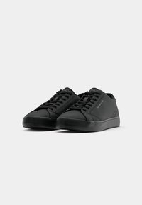 Paire de baskets basses noires avec lacets, marquage discret sur la languette et le côté, sur un fond clair uni.