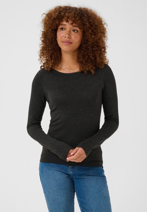 Femme aux cheveux bouclés portant une chemise sombre à manches longues et un jean bleu, debout, les mains jointes, regardant légèrement sur le côté.