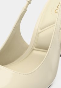 Chaussure à talons hauts beige avec une finition brillante, côtés ouverts et forme incurvée. Présente un design élégant et un intérieur rembourré.