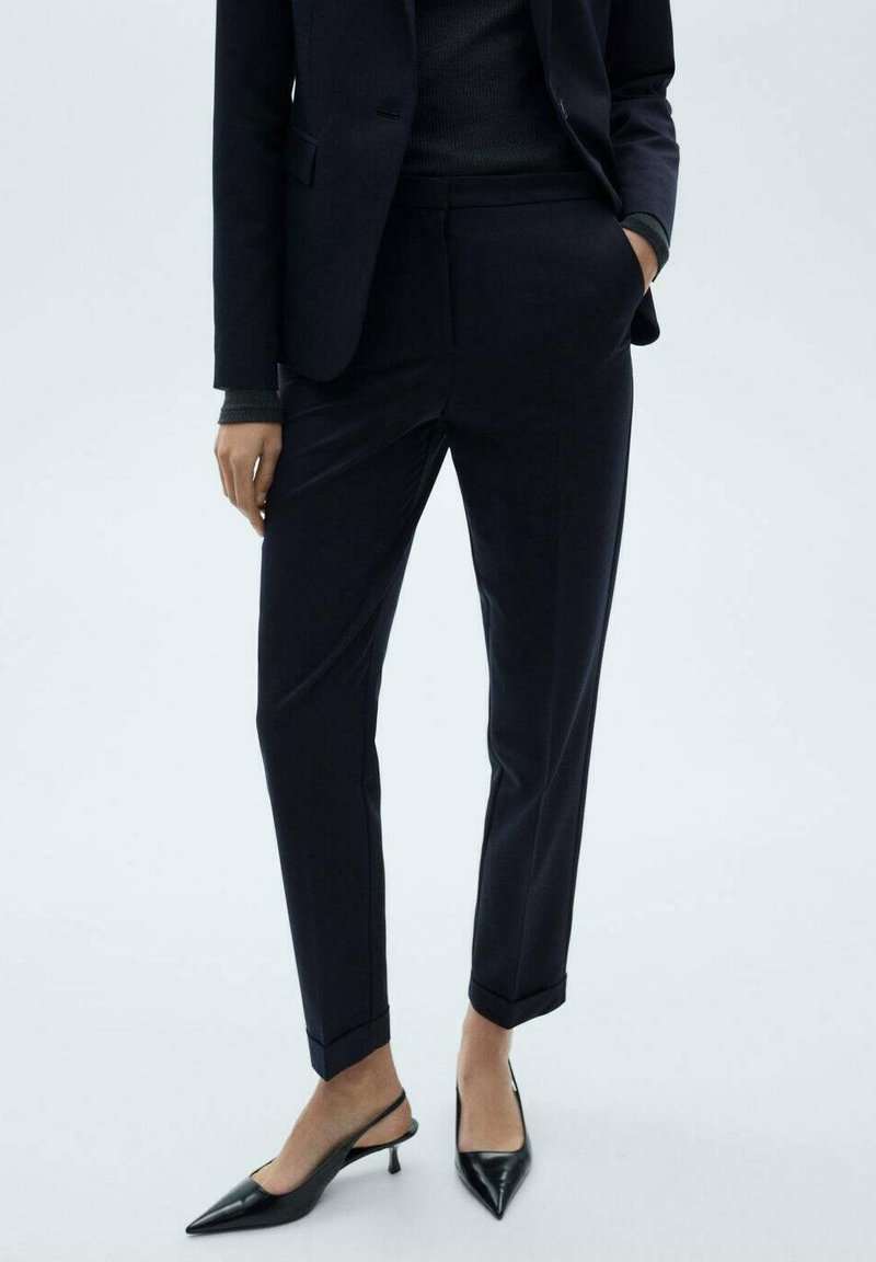 Pantalons tailleur bleu marine avec une coupe slim, présentant un devant plis, des poches latérales et une jambe fuselée. Associés à des escarpins noirs à bride arrière.