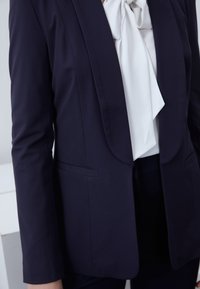 Veste bleu marine à coupe ajustée, col cranté et deux poches avant, associée à une blouse blanche à col lavallière en dessous. Texture de tissu lisse.