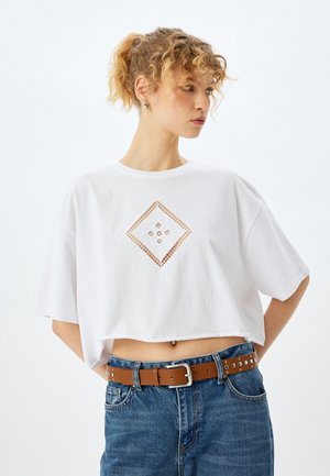 Witte cropped t-shirt van katoen, met een diamantvormige uitsnijding en kleine versieringen. Gekoeld met blauwe jeans en een bruine riem.